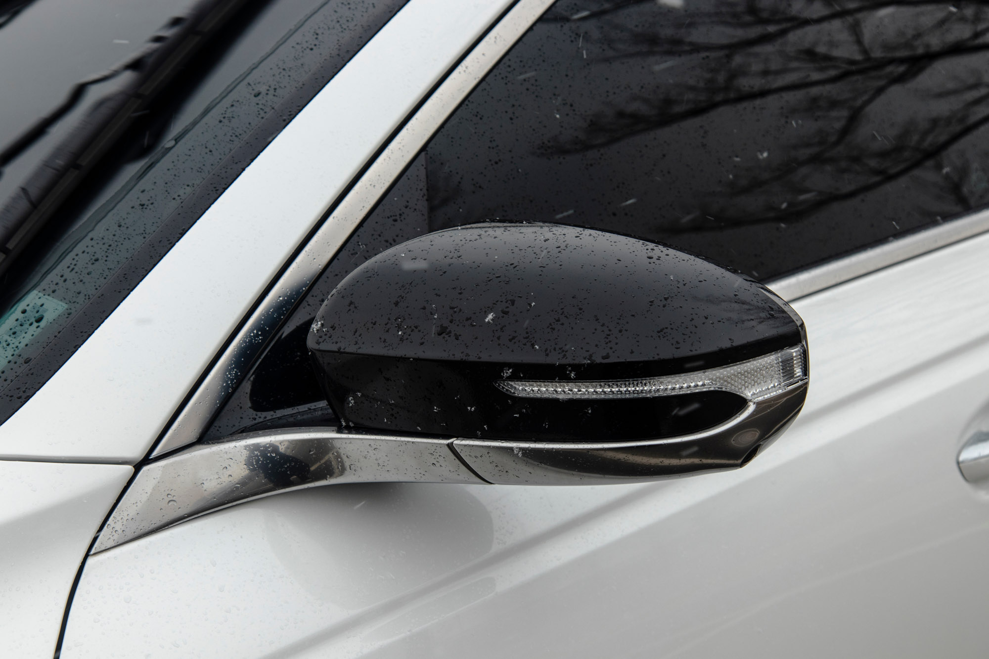 A close-up of a black side mirror on a white Kia K9 car, covered with small snowflakes and water droplets on a winter day.